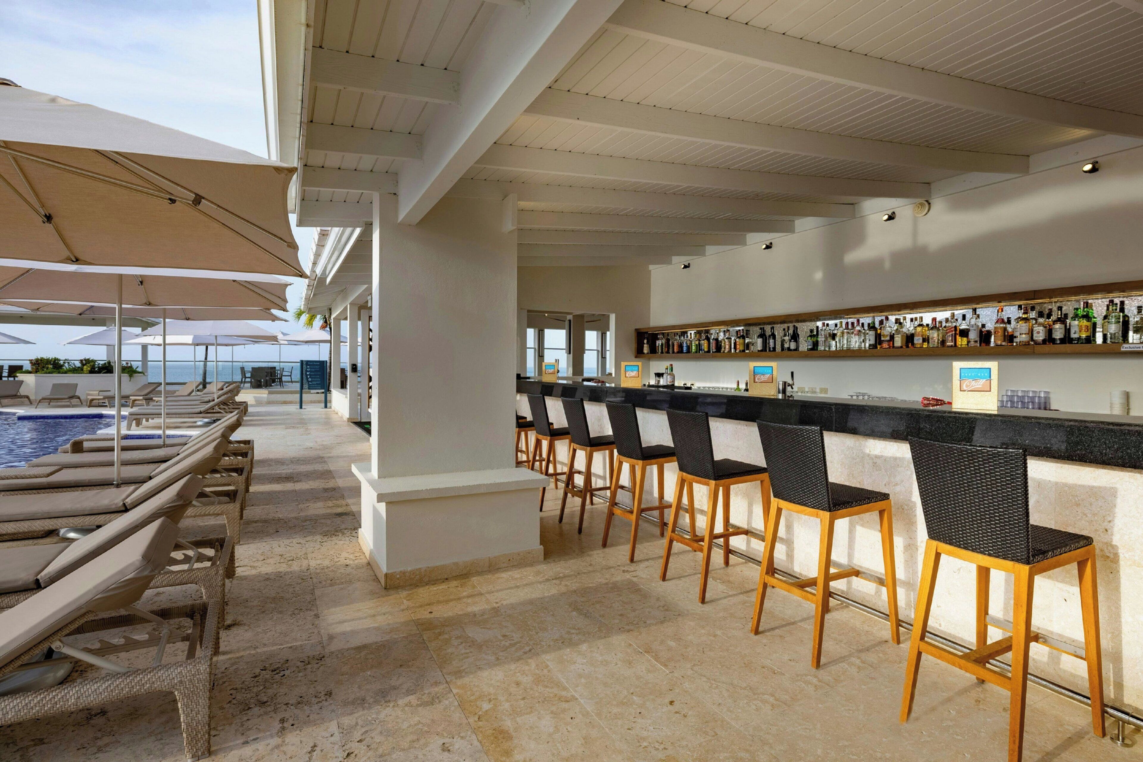 Zona de bar con vista a la piscina, con taburetes y una variedad de botellas en la estantería.