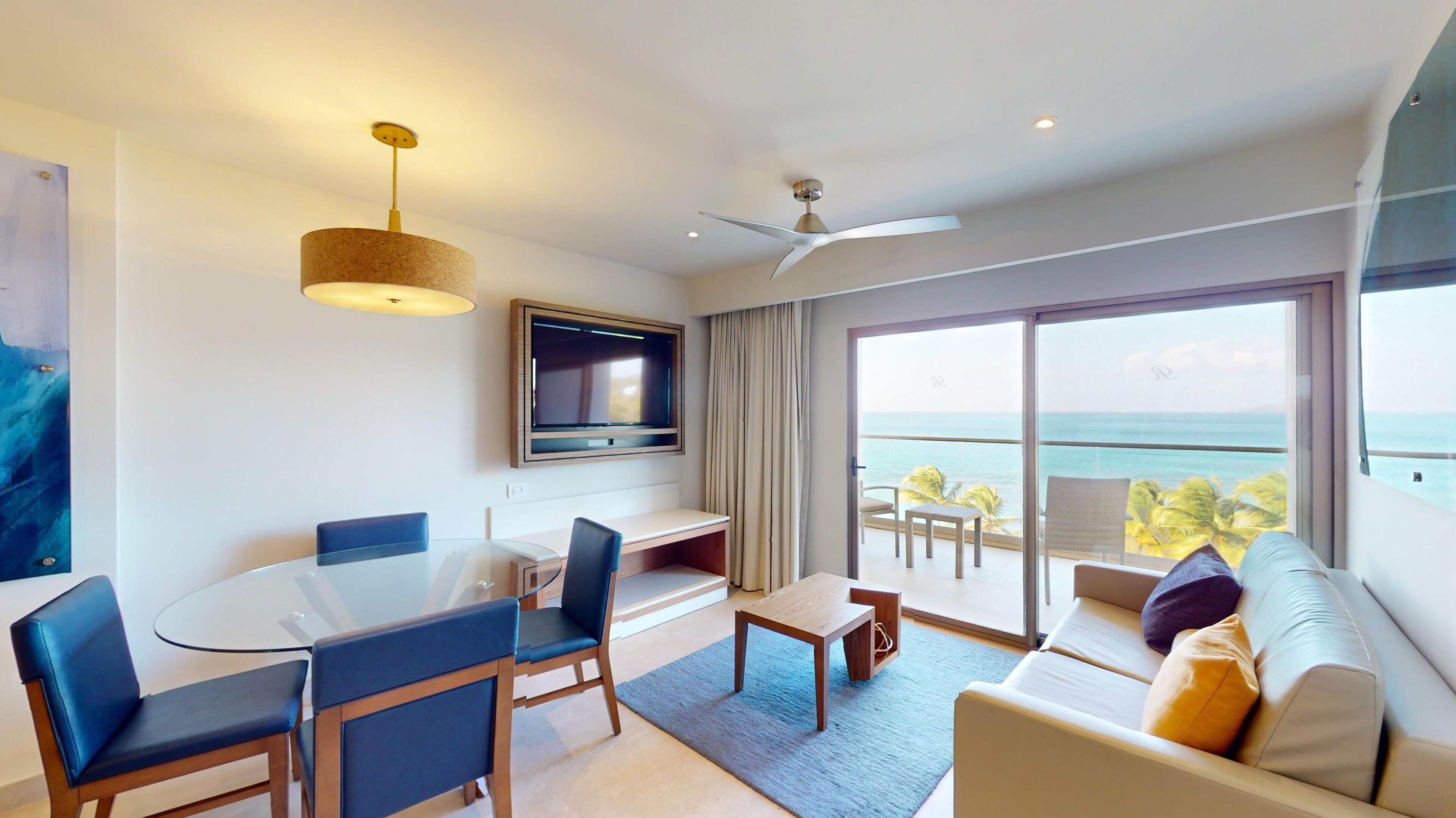 A modern hotel room with a dining area, a sofa, a TV, and a balcony with a view of the ocean.