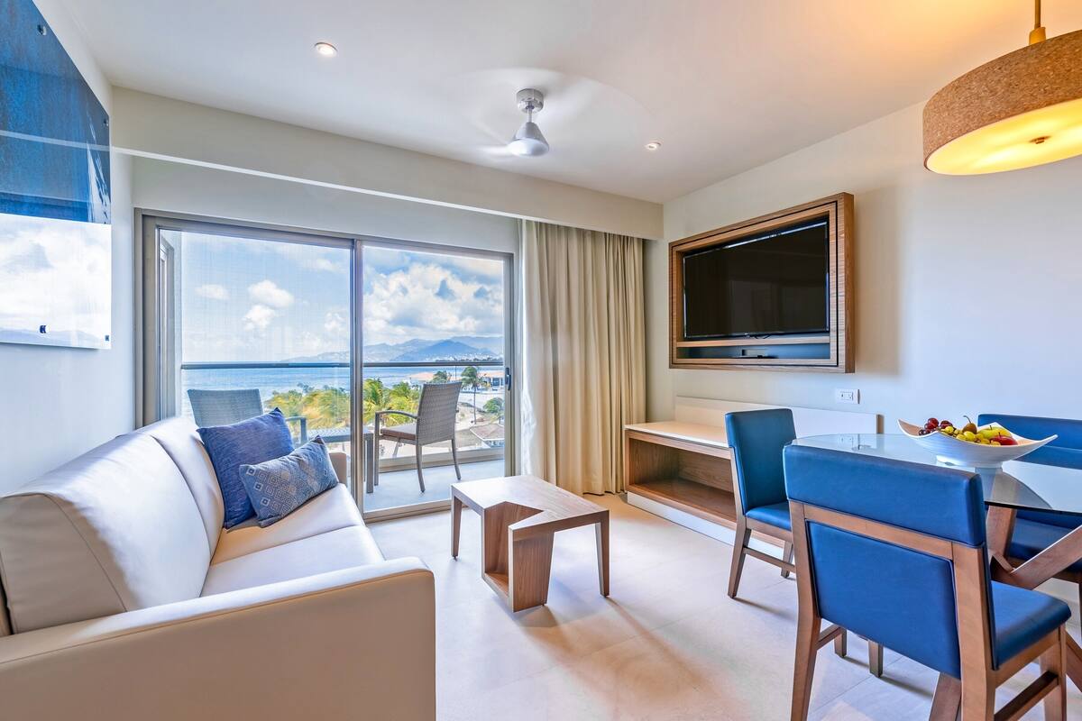 A modern living room with a sofa, coffee table, and dining area with chairs. A TV is mounted on the wall, and there is a large window with a view of the ocean.