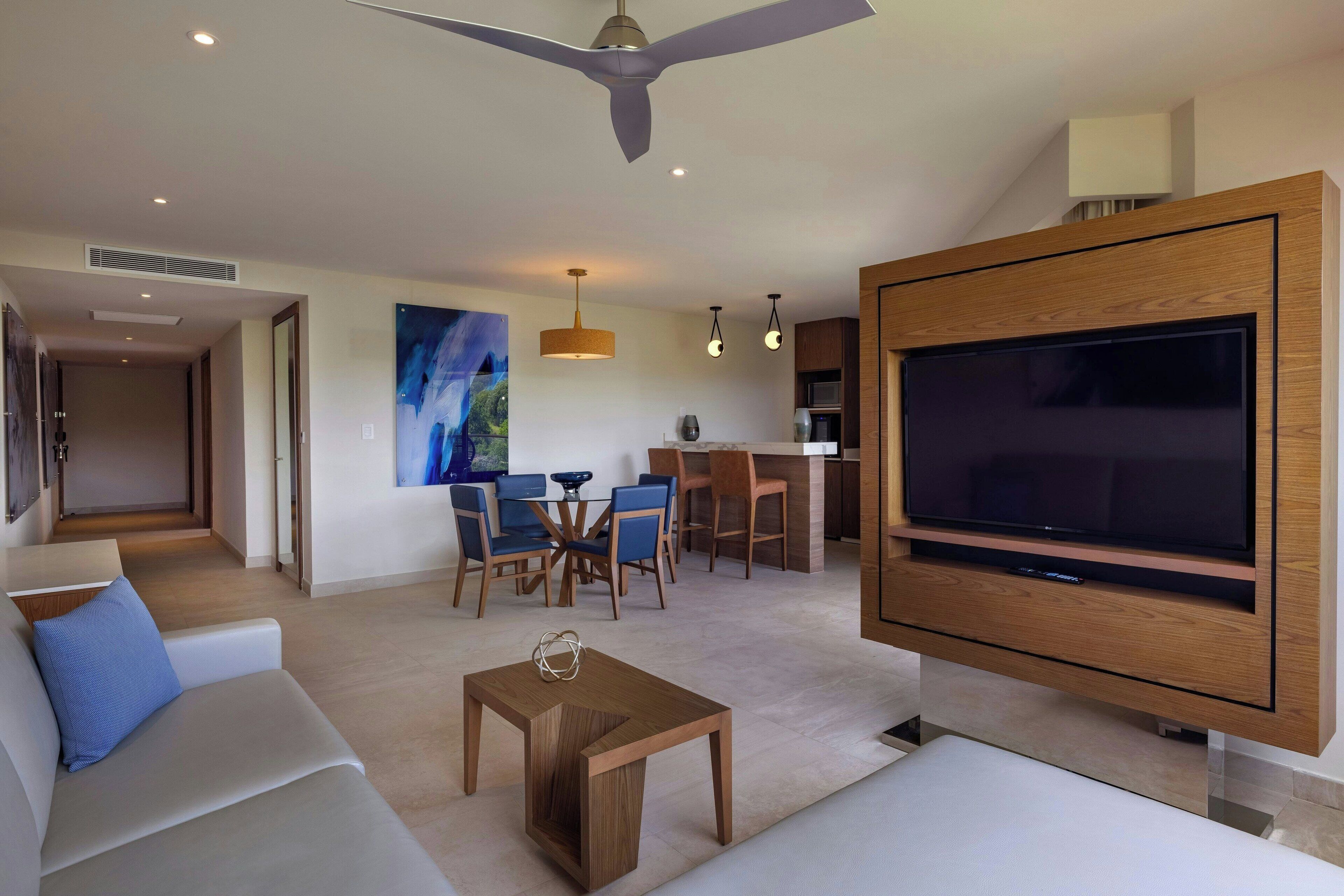 A modern living room with a large flat-screen TV, a ceiling fan, and a dining area with blue chairs.