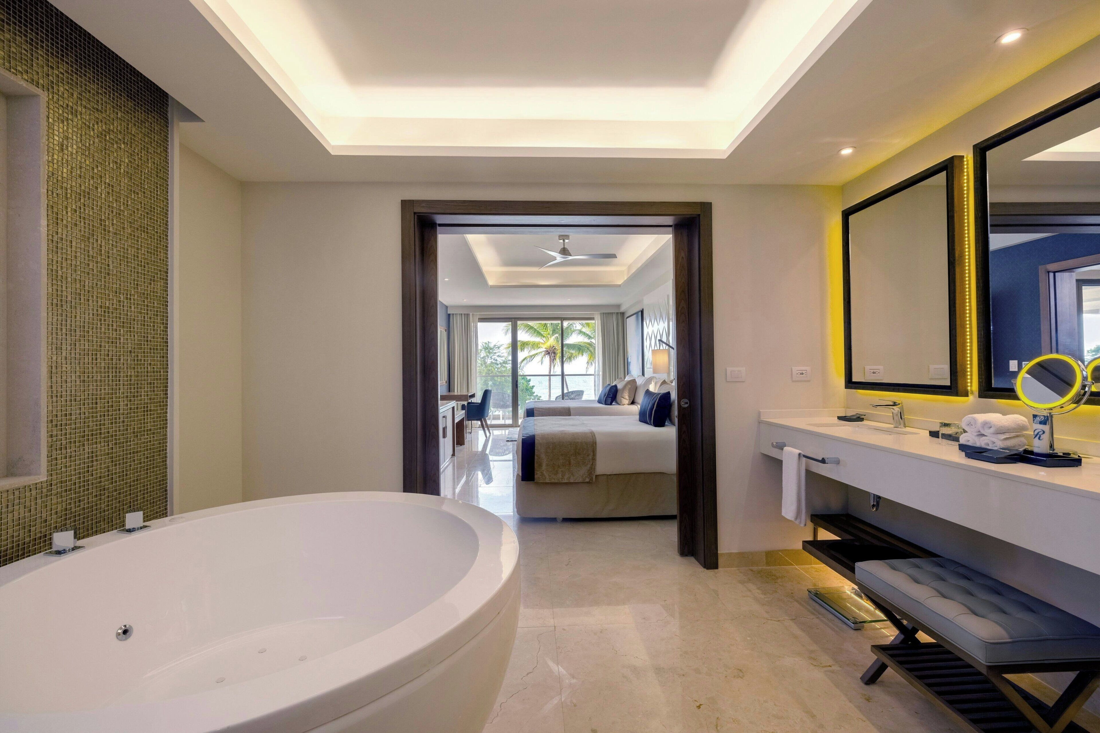 A modern bathroom with a large bathtub, a vanity with a mirror, and a view to a bedroom with a bed and a window.