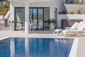 Un espace piscine extérieur moderne doté d’un patio fermé par des baies vitrées, de chaises longues et d’une table avec des chaises.