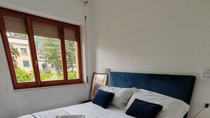 Siististi pedattu sänky, jossa on kuviollinen peitto ja tyynyjä, yöpöytä lampun kanssa ja ikkuna, josta avautuu näkymä rakennuksiin ja vihreään kasvillisuuteen.
