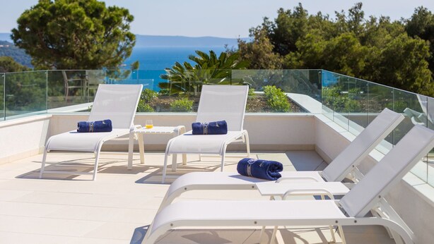 Une terrasse sur le toit, dotée de chaises longues blanches, d’une rambarde en verre et offrant une vue sur la mer et les arbres.