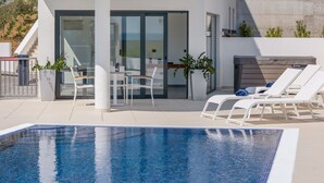 Un espace piscine extérieur moderne doté d’un patio fermé par des baies vitrées, de chaises longues et d’une table avec des chaises.