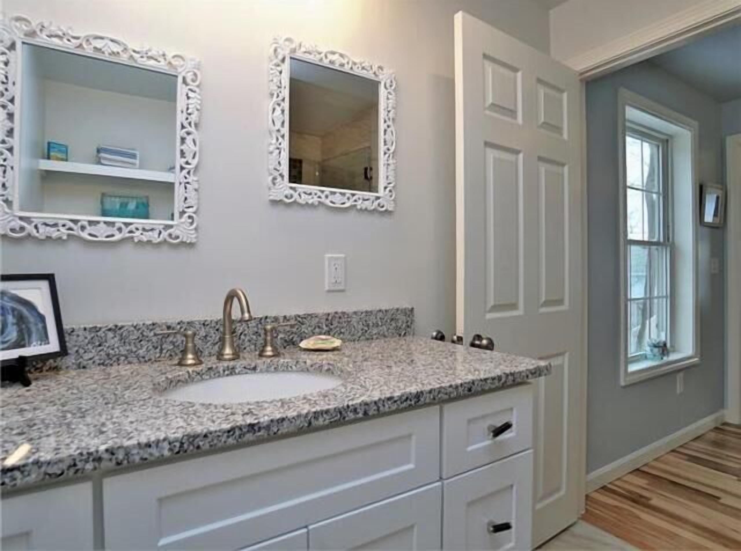 A bathroom with a granite countertop, a sink, two ornate mirrors, and white cabinetry.