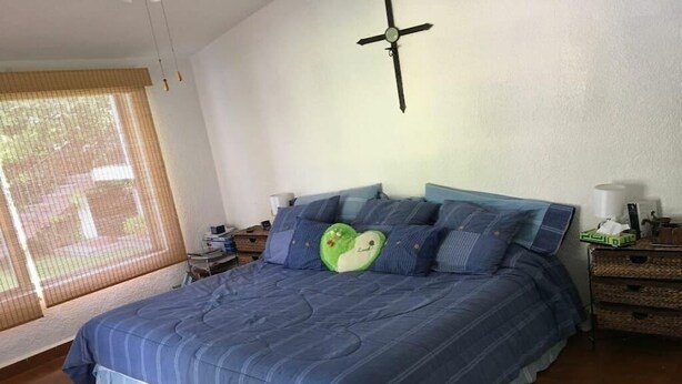 Et soveværelse med en stor seng, ventilator i loftet, et vindue med skodder, og et natbord med lampe og en grøn genstand.
