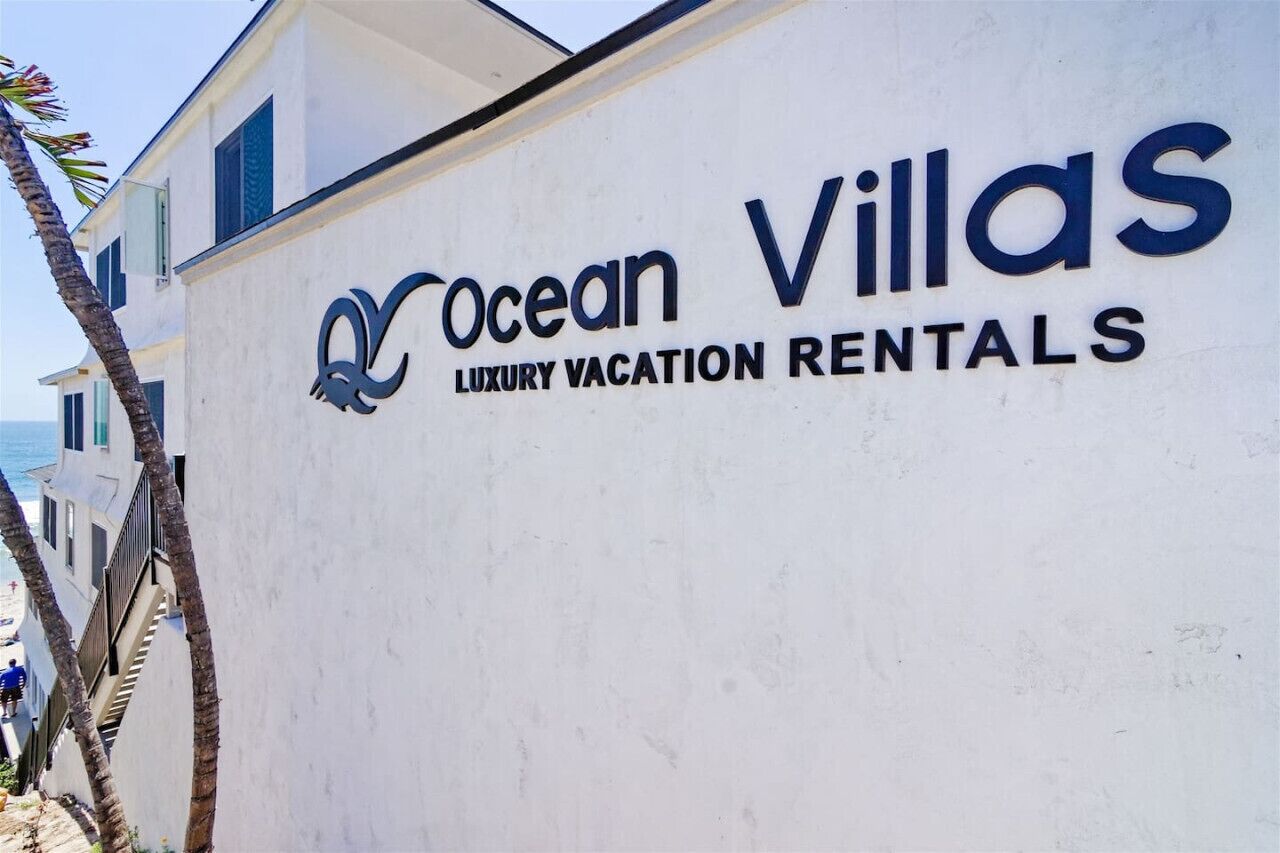 A sign for Ocean Villas Luxury Vacation Rentals with a clear view of the beach and ocean in the background.