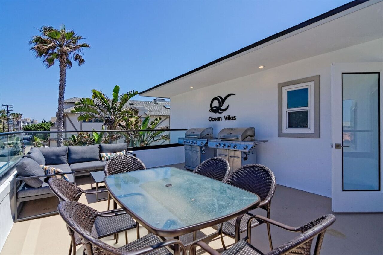 Una terraza en la azotea con barandilla de cristal, mobiliario exterior, una parrilla y una palmera.