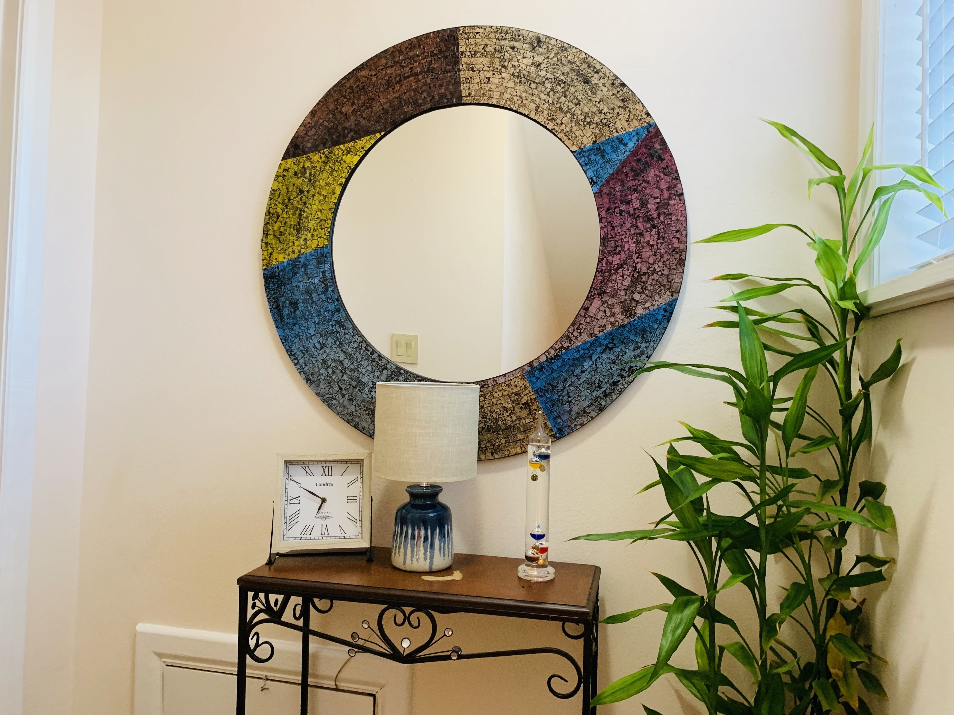 A wall-mounted circular artwork with a clock and a lamp on a table beside it.