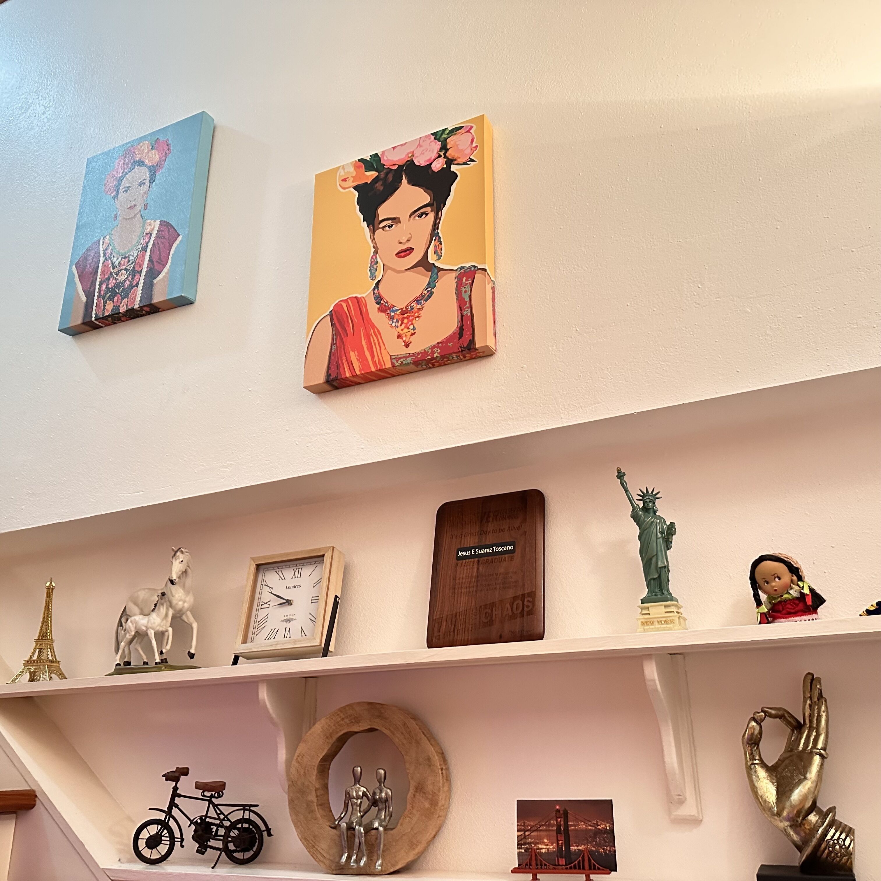 Een muur met twee schilderijen, een klok, een plaquette, een beeldhouwwerk, een fiets, een houten frame met figuren en een gouden sculptuur van een hand.