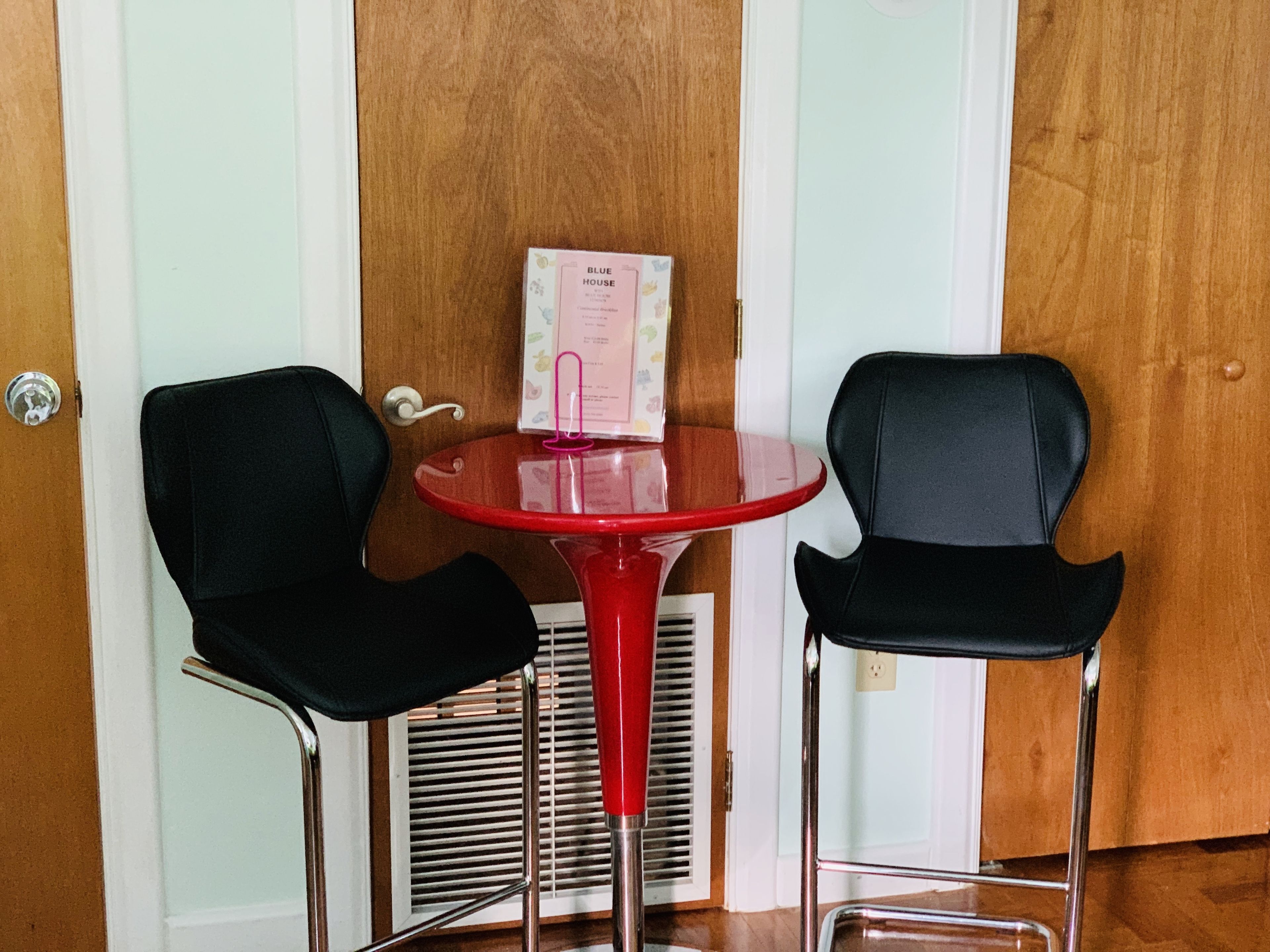 Une petite pièce avec une table rouge et deux chaises noires, une porte en bois et un mur blanc.