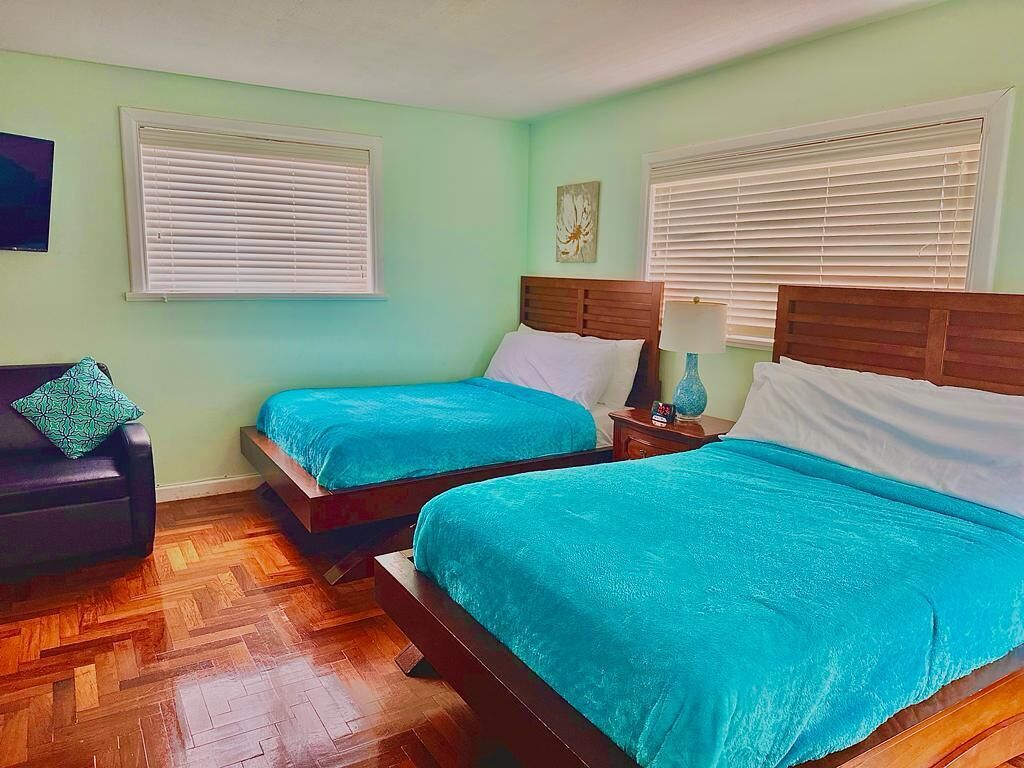 Ein Zimmer mit zwei Betten, jeweils mit einem Holz-Kopfteil und türkisblau gewaschener Bettwäsche. Es gibt einen Holzboden und ein dunkelgraues Sofa mit einem gemusterten Kissen.