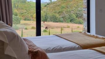 Ett hotellrum med ett stort fönster som erbjuder utsikt över ett kuperat landskap med gles vegetation och ett staket.