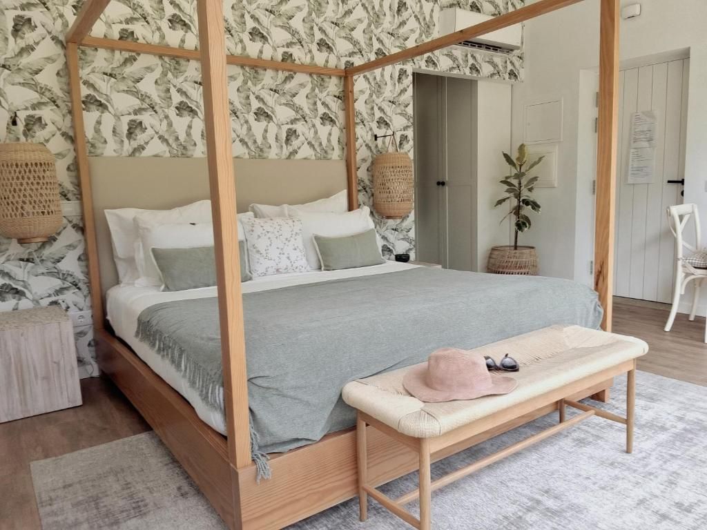 Una cama con dosel con cubrecama gris, un banco con sombrero y gafas de sol, y una pared con estampado de hojas.