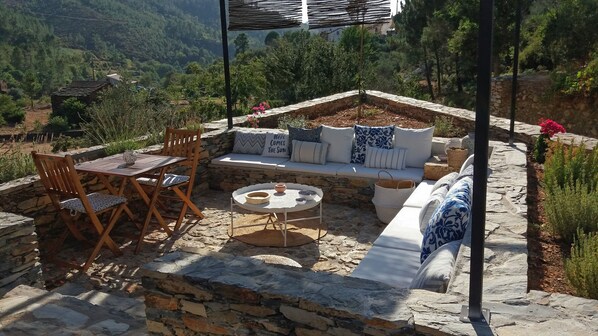 A stone patio with a wooden table and chairs, a round table with a bowl, a stone bench with cushions, and a view of the surrounding landscape. - Quinta das Levadas Country House & Nature (Oliveira do Hospital)