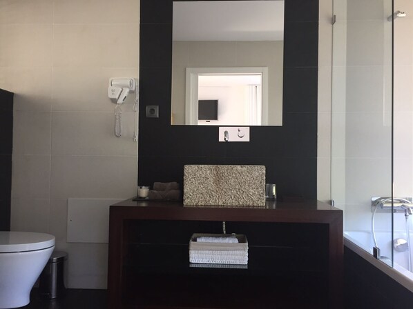 A modern bathroom with a dark tiled wall, a white toilet, a wooden vanity with a granite countertop, a mirror, and a glass shower enclosure.