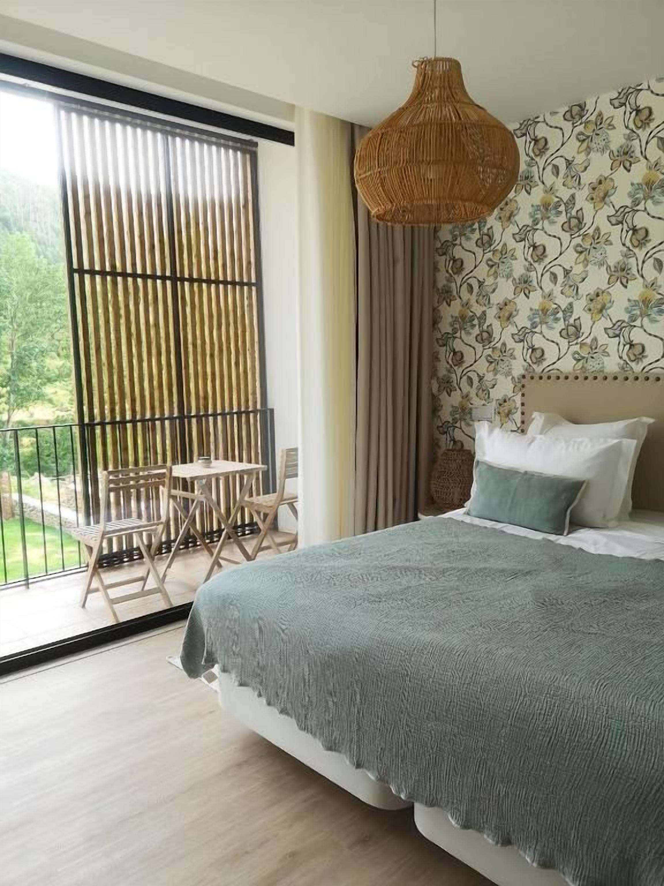 Ein Schlafzimmer mit Bett, einer Pendelleuchte, einem Fenster mit Blick und einem Balkon mit Tisch und Stühlen.