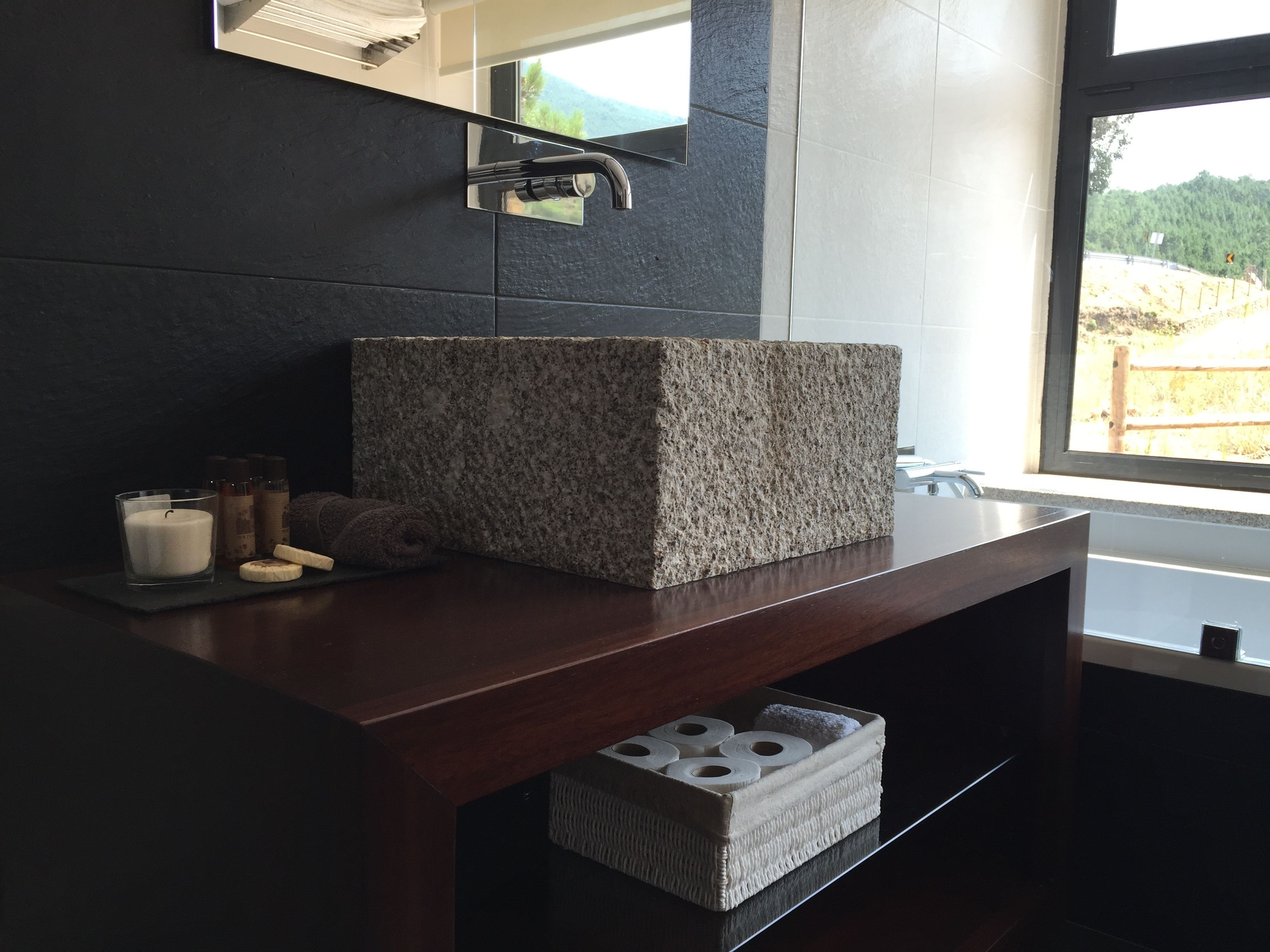 A bathroom with a granite sink, a mirror, a candle, and a window with a view of a forest.