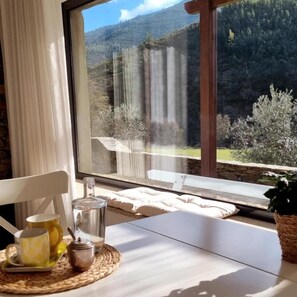 Ein Zimmer mit einem großen Fenster mit Blick auf eine gebirgige Landschaft, einem Tisch mit einer Karaffe und Tassen sowie einem Stuhl.
