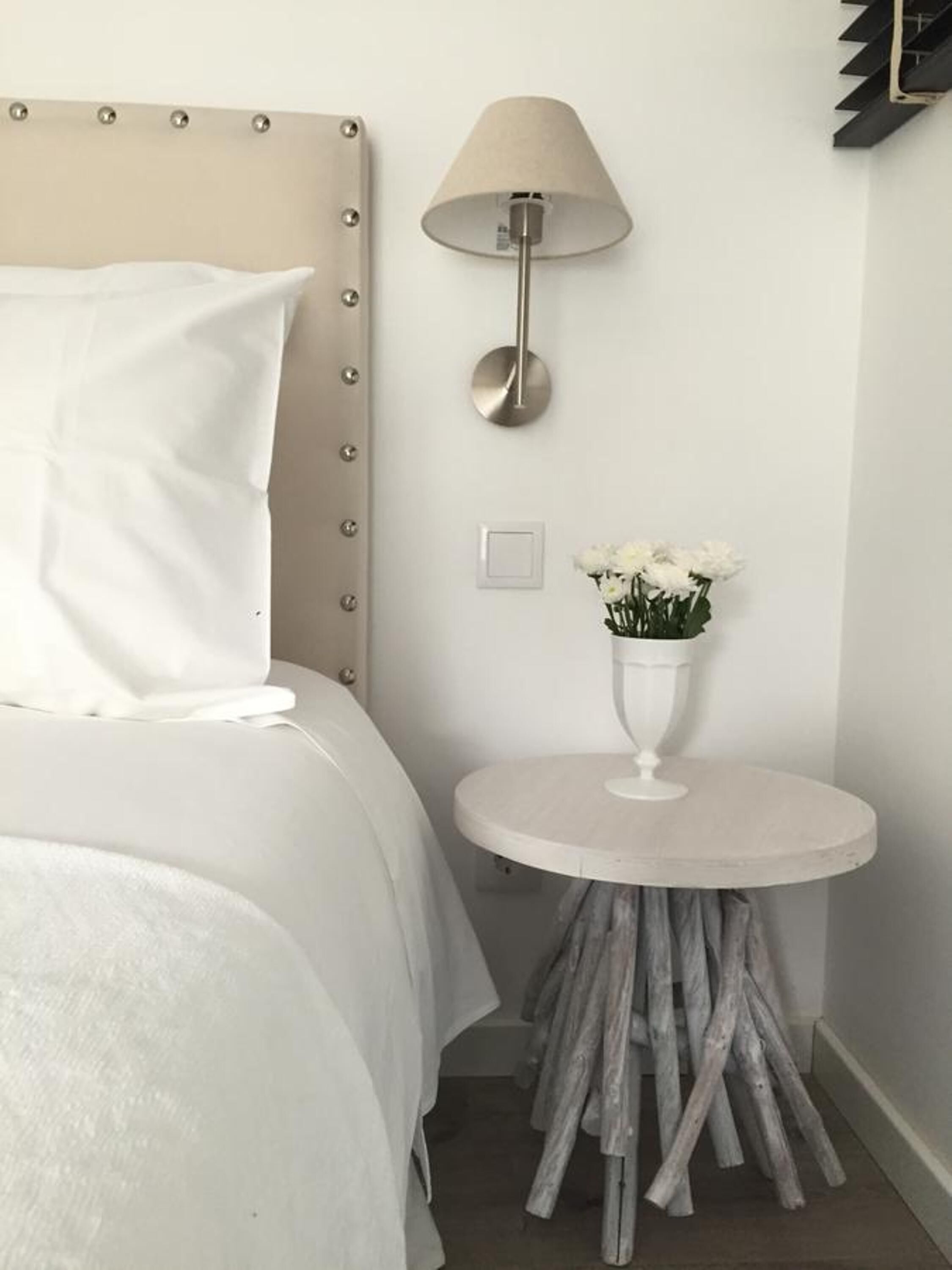 Una cama bien tendida con sábanas blancas, una lámpara de pared y una pequeña mesa redonda con un florero de flores blancas.