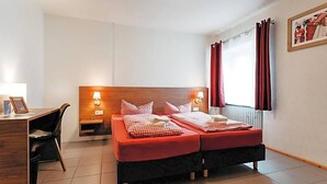 A hotel room with a wooden headboard, two beds with red bedding, a desk with a chair, and a window with red curtains.