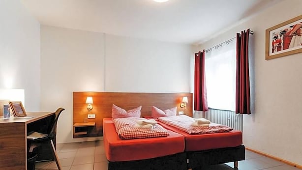 A hotel room with a wooden headboard, two beds with red bedding, a desk with a chair, and a window with red curtains.