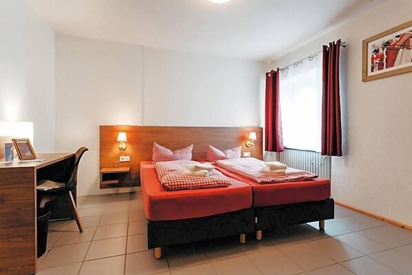 A hotel room with a wooden headboard, two beds with red bedding, a desk with a chair, and a window with red curtains.