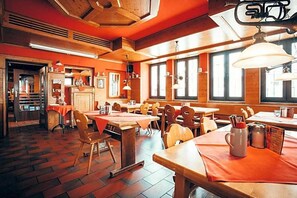 A restaurant interior with wooden tables and chairs, large windows, and a brick floor.