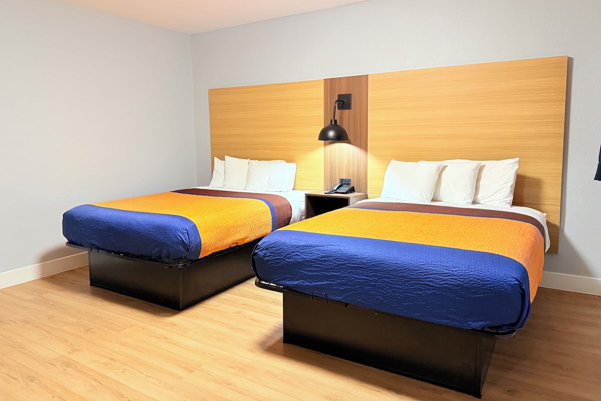 Habitación de hotel con dos camas, cabecero de madera, mesitas de noche y una mesita de noche con lámpara.