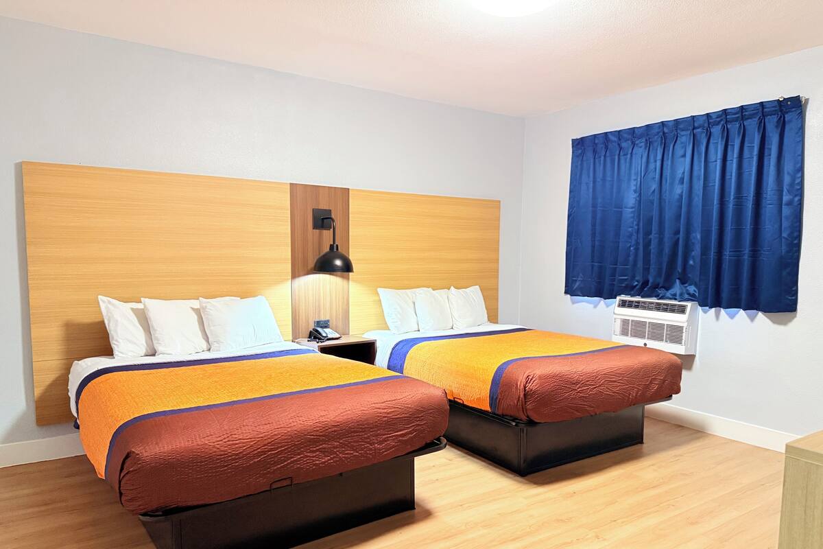 Habitación de hotel con dos camas, cabecero de madera, mesita de noche, lámpara, ventana con cortinas azules y unidad de aire acondicionado.
