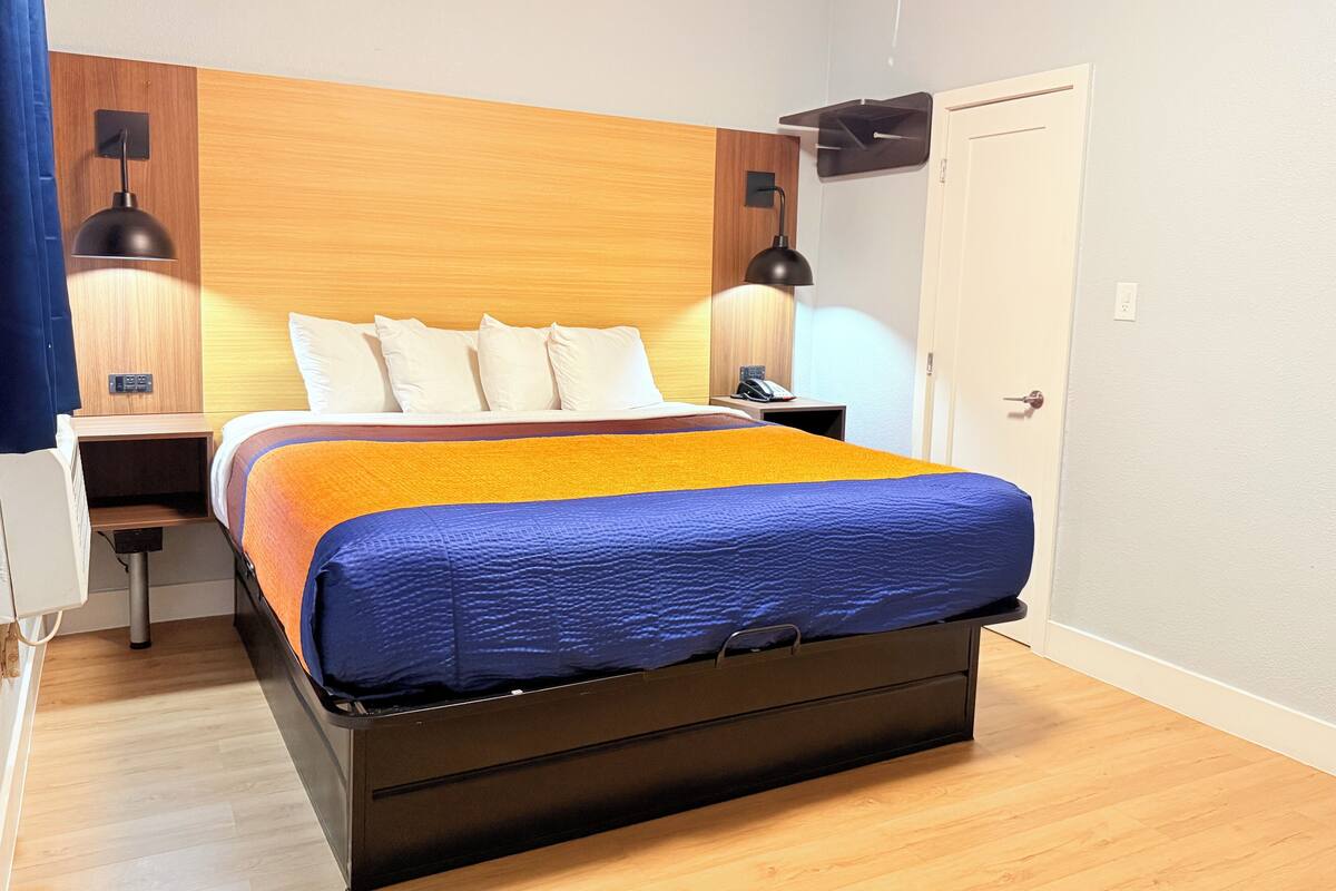Una habitación de hotel moderna con cabecera de madera, una cama con edredón azul y naranja, dos mesitas de noche con lámparas y una puerta.