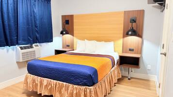 Quarto de hotel com cabeceira de madeira, cama com colcha azul e laranja, duas luminárias de apoio, janela com cortinas azuis e um ar condicionado.