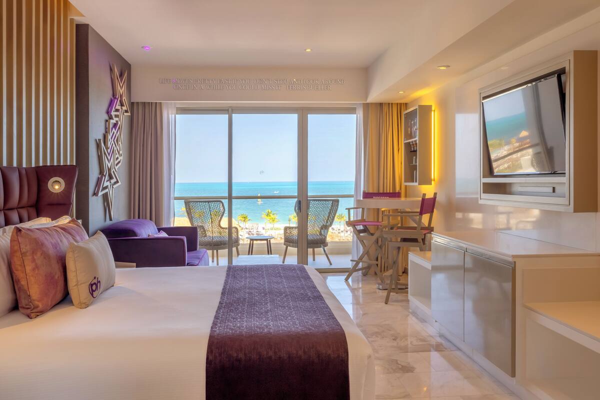 A hotel room with a large bed, a TV, a desk, and a view of the ocean.