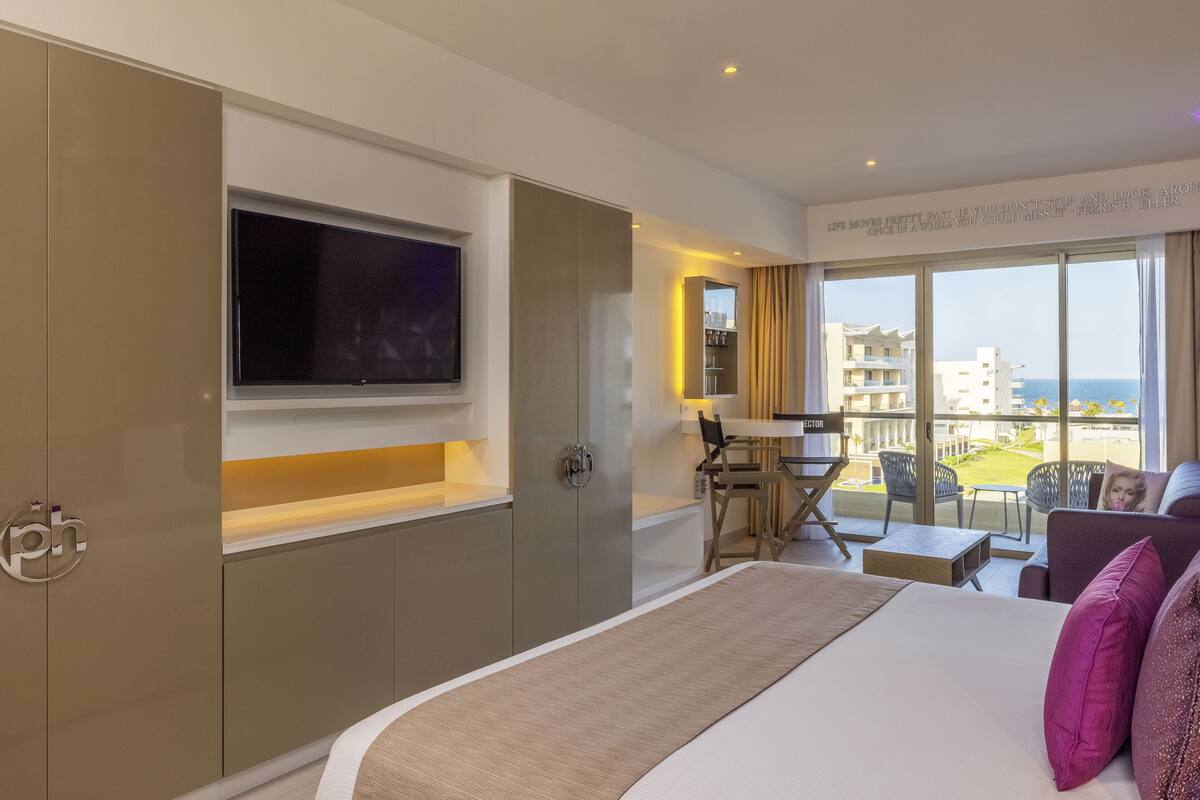 A hotel room with a large bed, a TV mounted on the wall, a desk with a chair, and a balcony with a view of the sea.