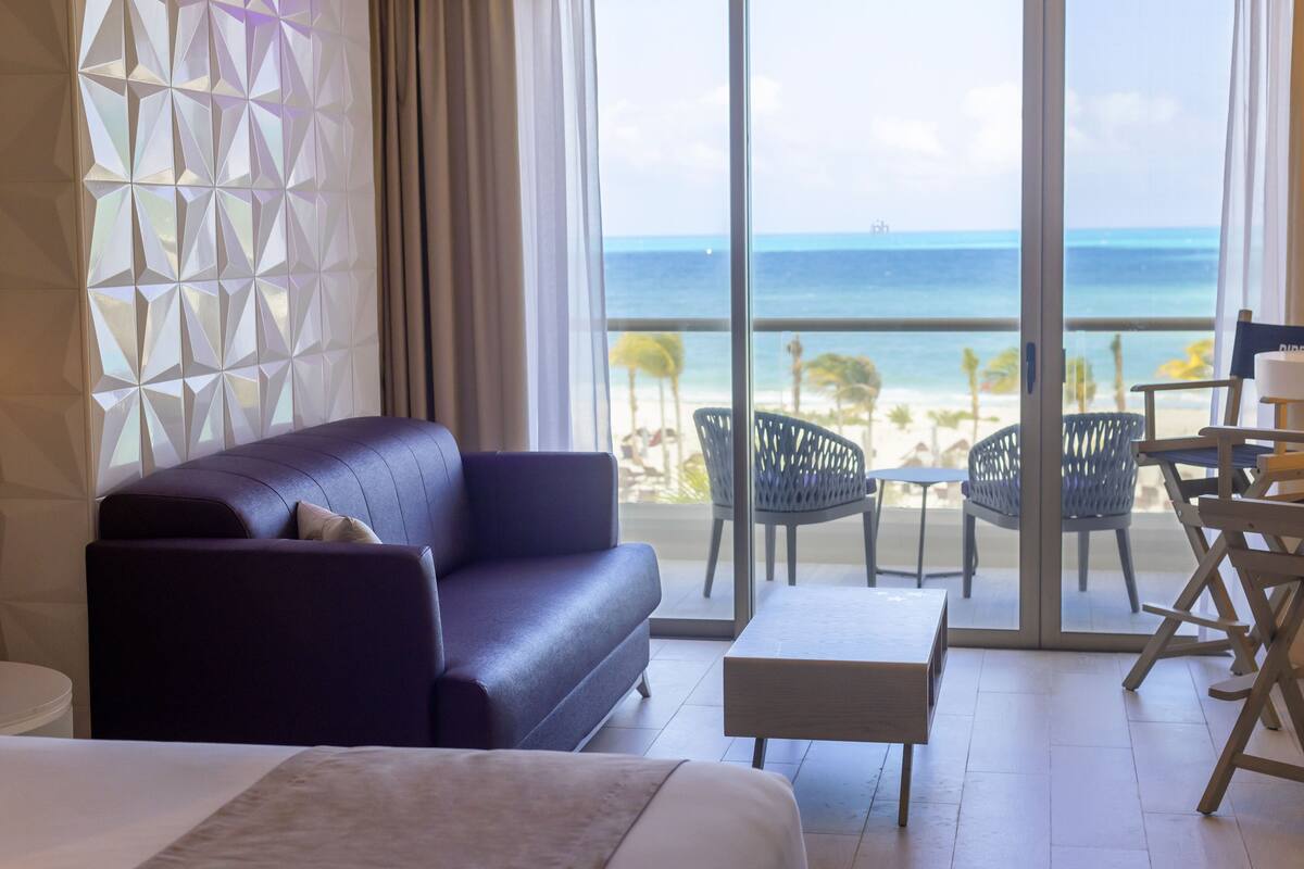 A room with a large bed, a sofa, a small table, and a balcony with a view of the beach and ocean.