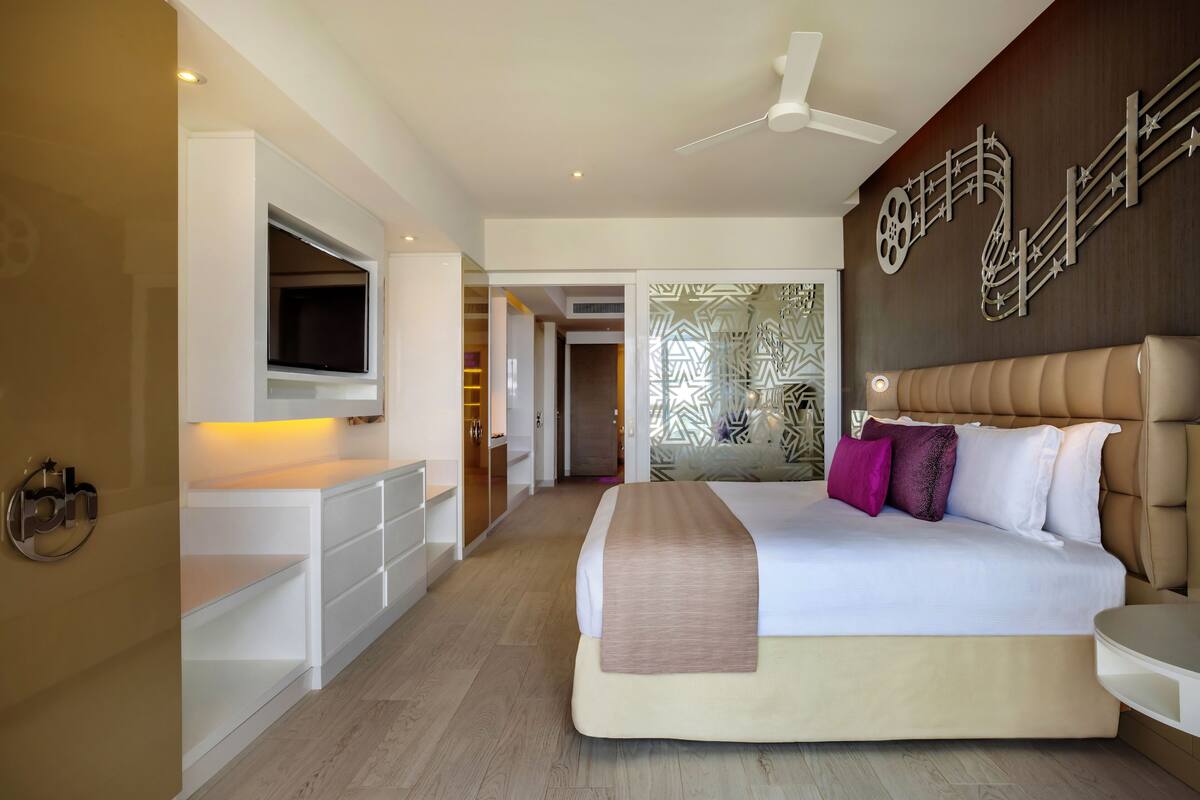 A modern hotel room with a large bed, a flat-screen TV mounted on the wall, a ceiling fan, and a decorative wall piece.