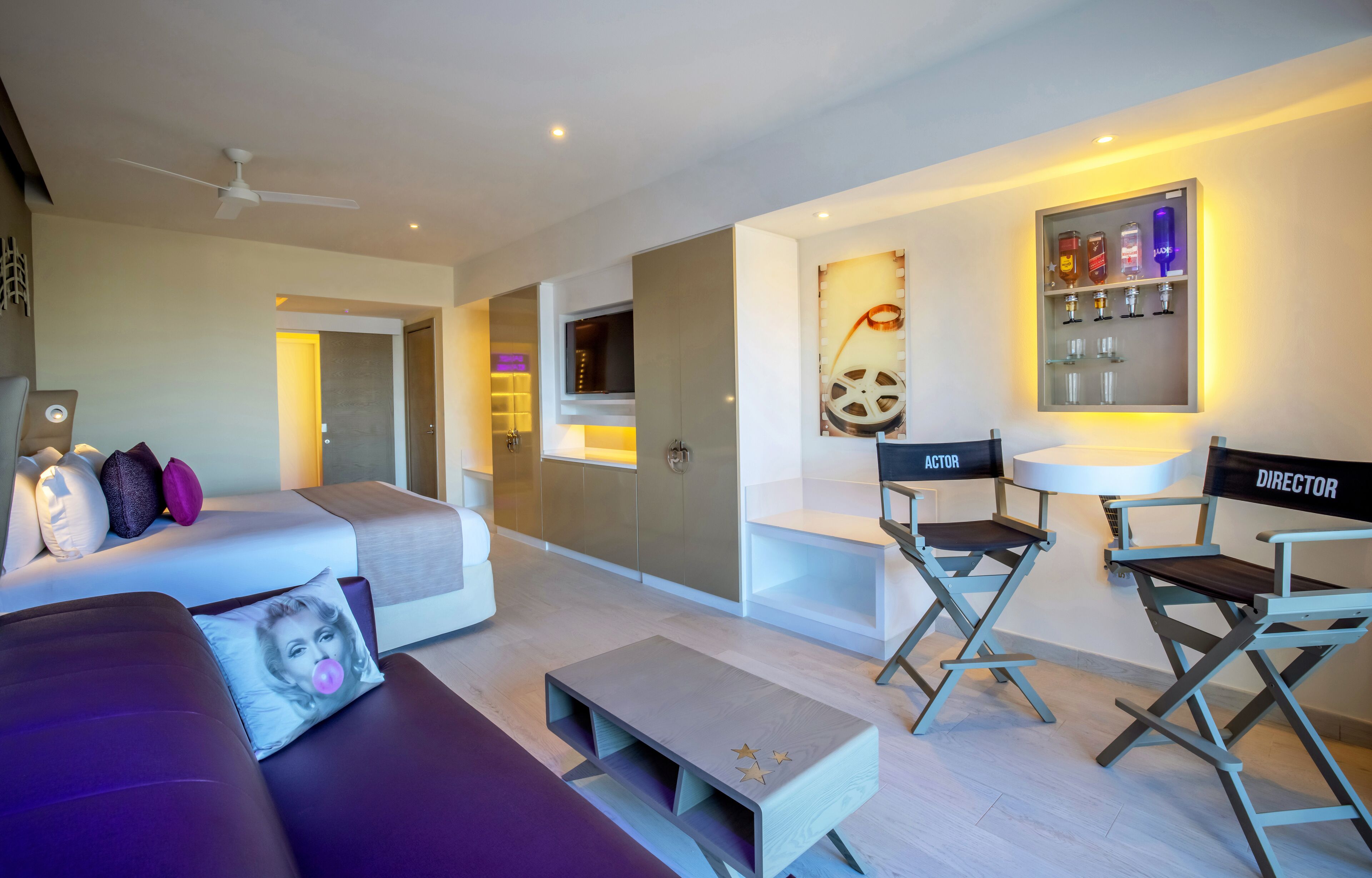 A modern living room with a sofa, coffee table, and director chairs. There is a TV, a cabinet, and a wall-mounted shelf with glasses and bottles.