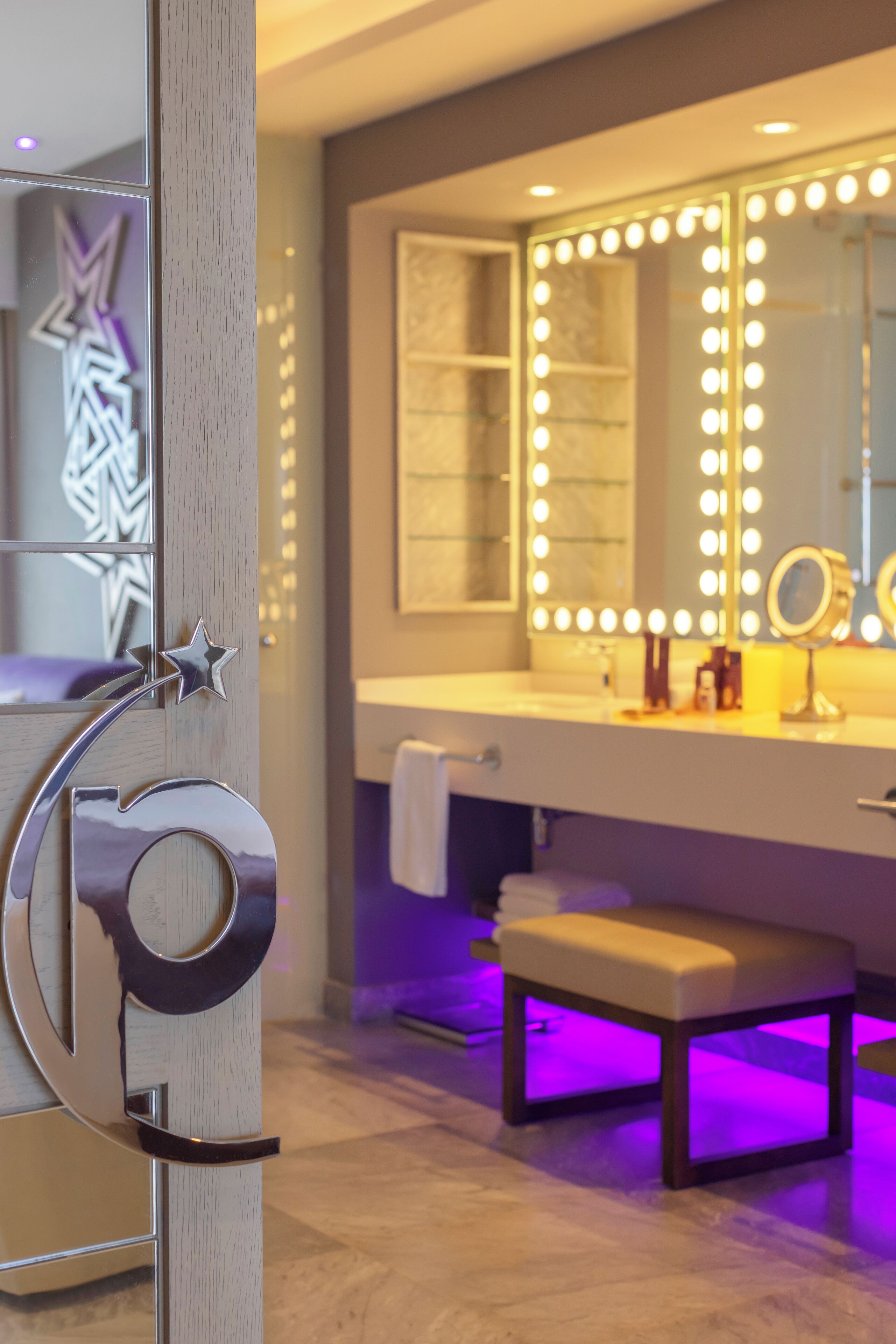 A modern bathroom with a vanity, a large mirror with lights, a bench, and a star-shaped decoration.