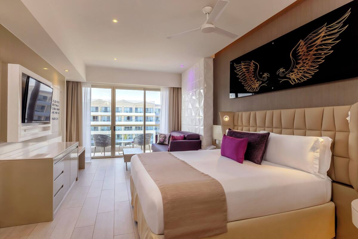 A modern hotel room with a large bed, a TV, a desk, and a balcony with a view of the city.