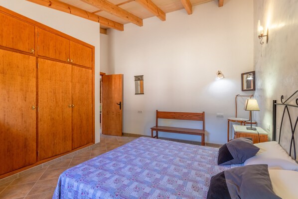 Une chambre à coucher comprenant un lit, une armoire en bois, un banc, une table de chevet, une lampe et un luminaire fixé au mur.