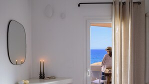 Una stanza con uno scaffale bianco, uno sgabello di legno, uno specchio e una porta che dà su un balcone con vista sul mare.