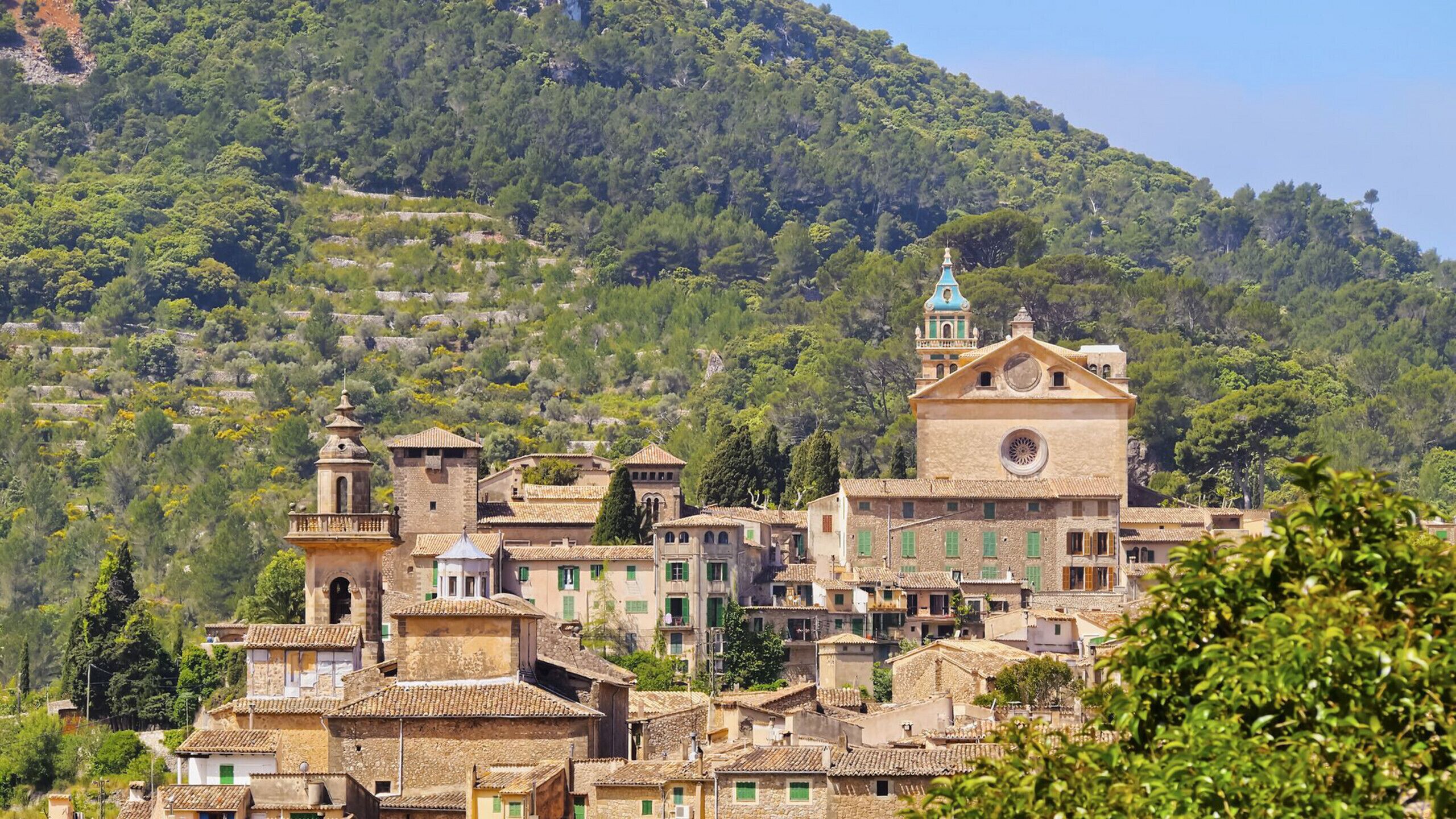 A historic town with terracotta-roofed buildings, a prominent church with a bell tower, and a hillside backdrop.