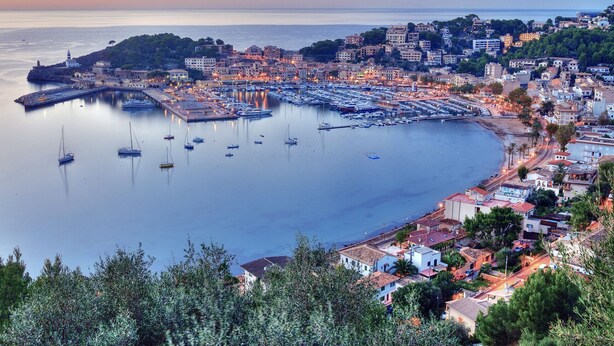 Un pueblo costero con un puerto deportivo, edificios y un paisaje montañoso de fondo.