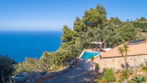 Zona de piscina con vista al mar, rodeada de árboles y un muro de piedra.
