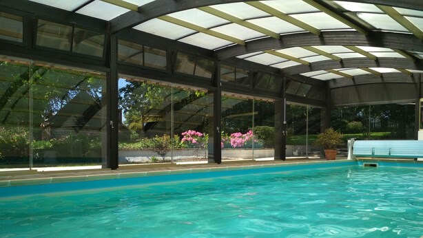A swimming pool with a glass enclosure and a covered roof, surrounded by potted plants and flowers. - Near Baie de Somme "good authie" (Colline-Beaumont)