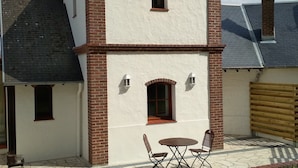 A two-story building with a white exterior and red brick accents, featuring a small patio with a table and chairs. - Near Baie de Somme "good authie" (Colline-Beaumont)