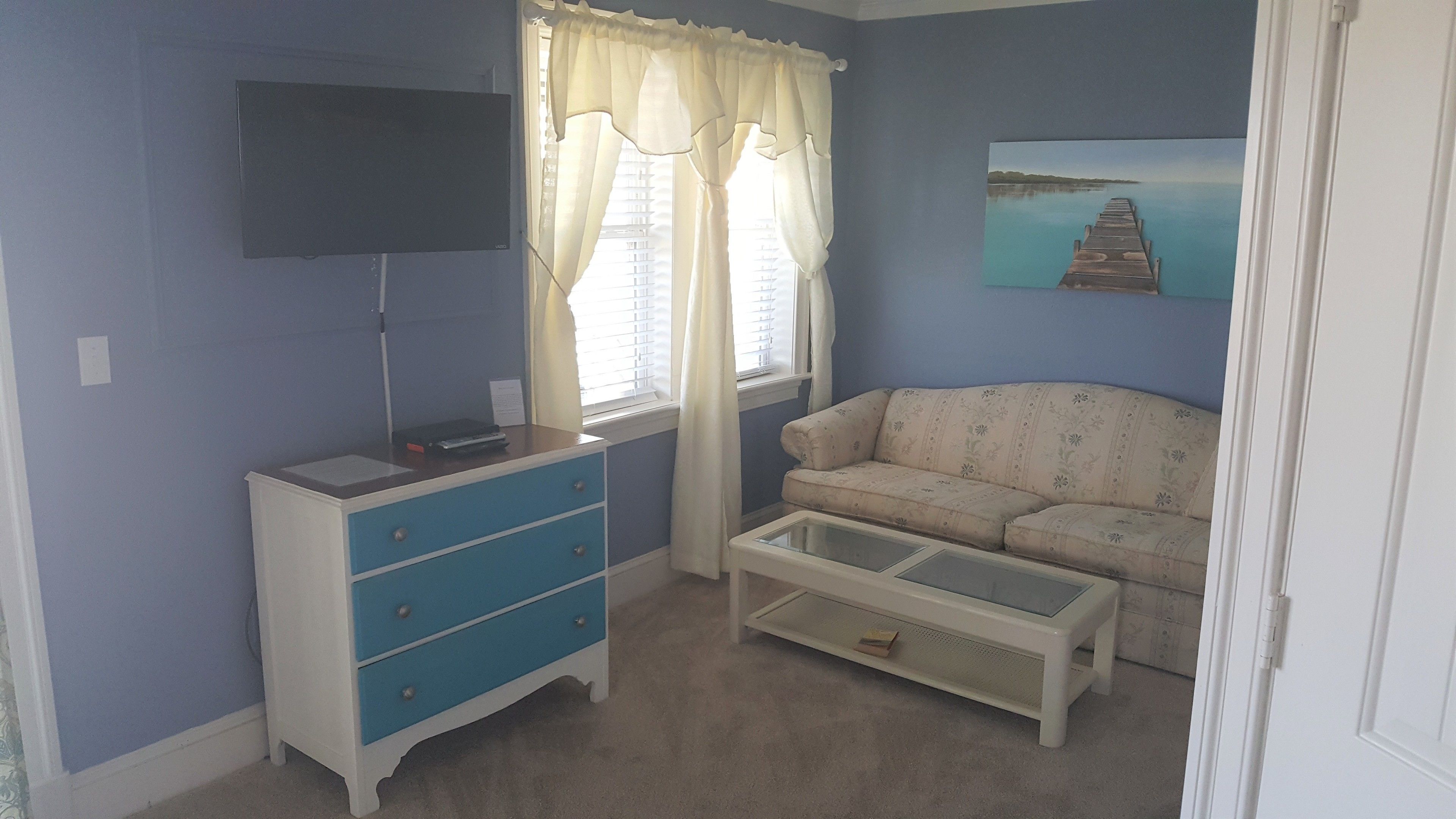 A room with a blue wall, a white sofa, a small table, a TV on the wall, and a window with curtains.