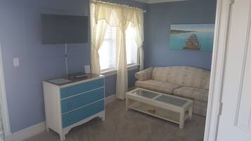 A room with a blue wall, a white sofa, a small table, a TV on the wall, and a window with curtains.