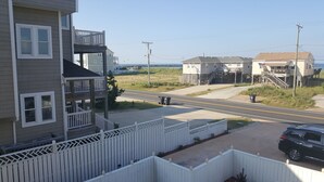 A residential area with a two-story house featuring a balcony, a street with a parked SUV, and a clear view of the beach in the background. - Pierhouse Bed & Breakfast (Kill Devil Hills)